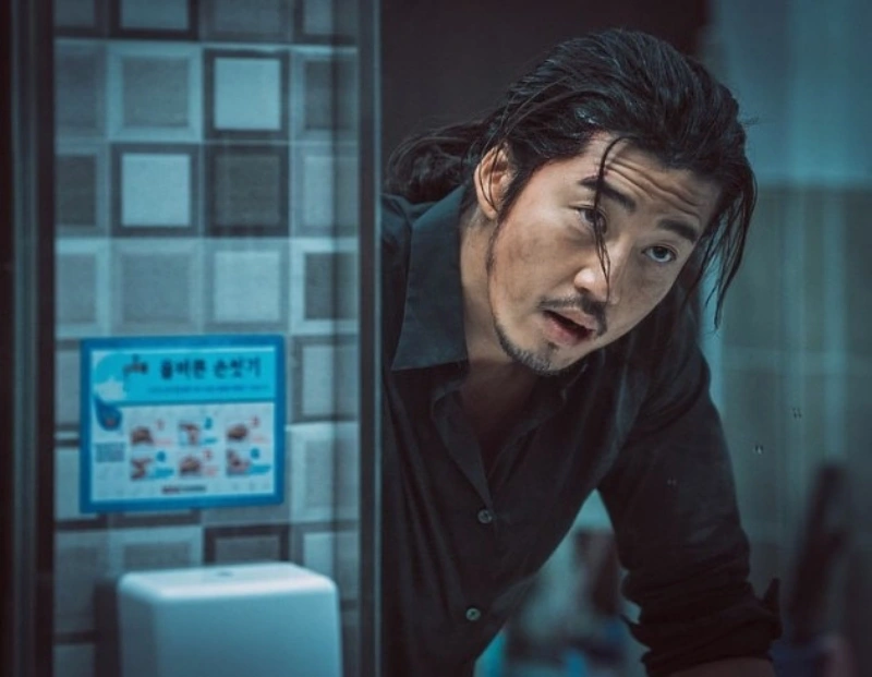 긴 머리와 검은 와이셔츠를 입고 화장실 세면대 거울을 보면서 서 있는 영화 범죄도시의 장첸 캐릭터